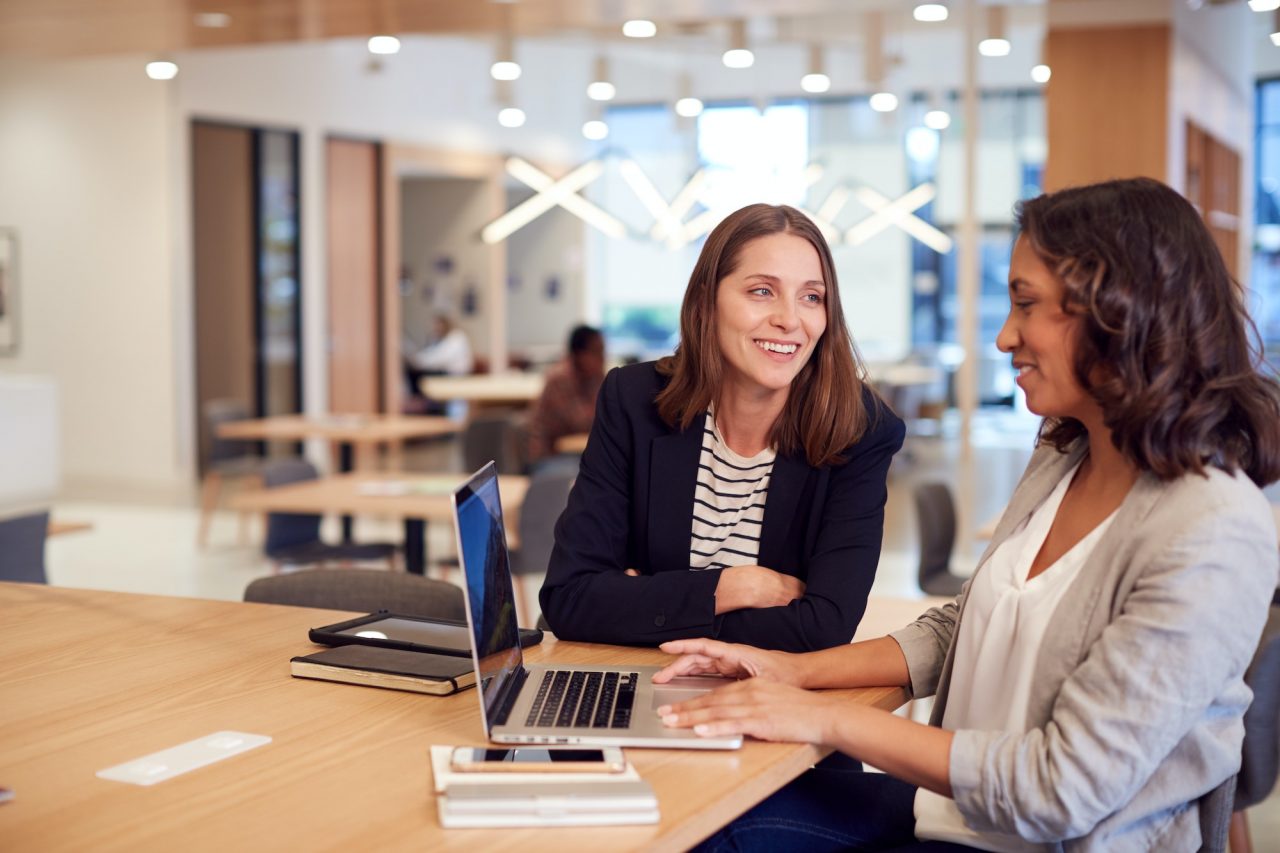 two businesswomen with laptop at desk in open plan office collaborating on project together 1
